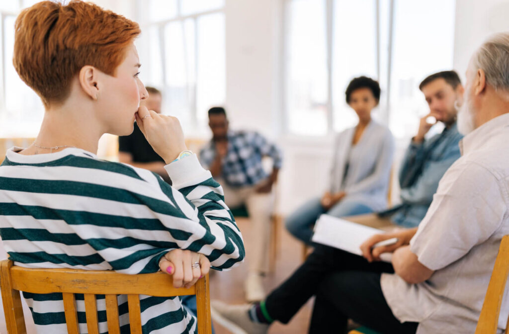 A group sitting in a circle during a therapy session in rehab, smiling and listening to their counselor speak.