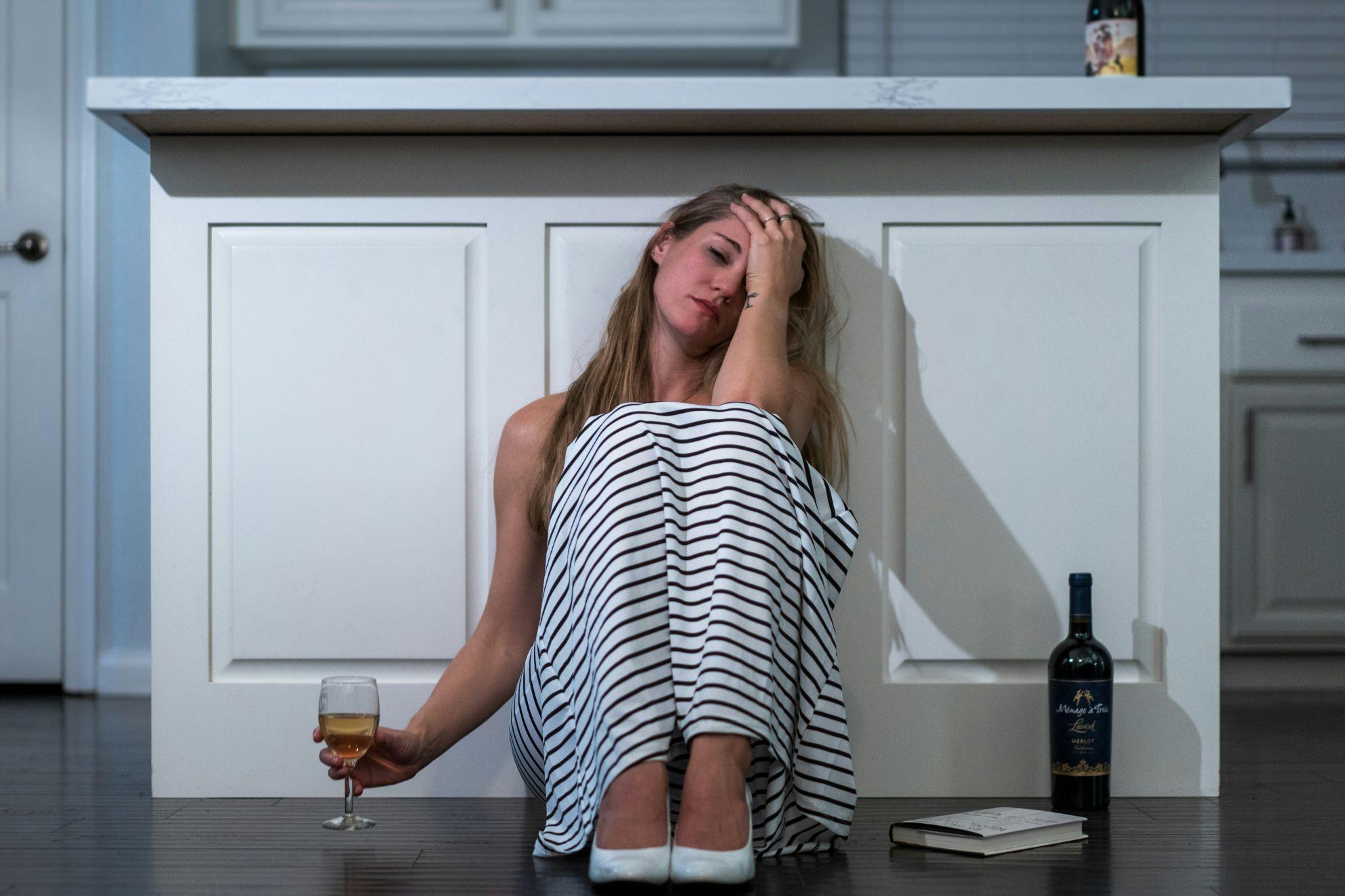 A young woman sitting in the kitchen with her hand on her face and holding a glass of wine, afraid of her appearance now being a part of the many faces of alcohol.