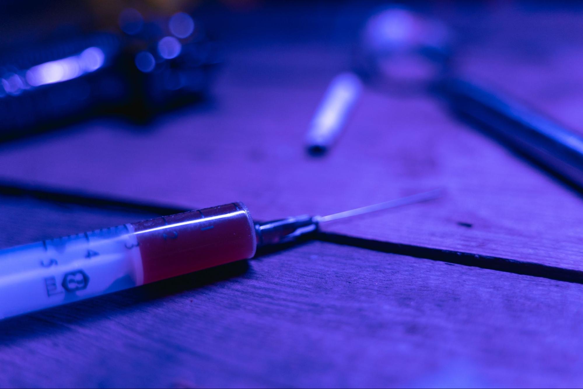 A needle and syringe lie on a table among other drug items, the culprit of track marks.