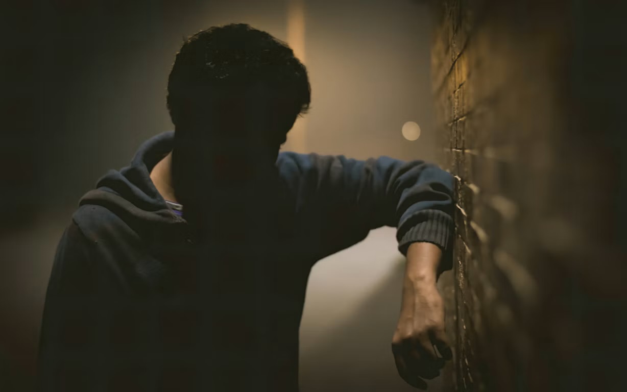 A man covered in shadow leaning against a brick wall, overgeneralizing his addiction recovery efforts.