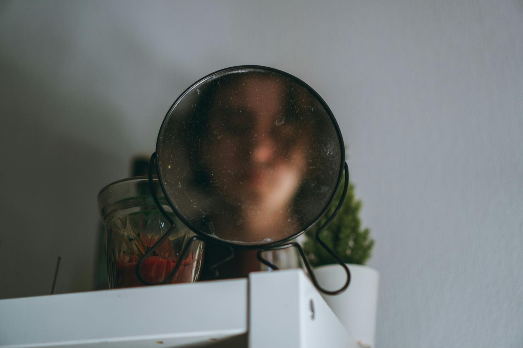 A person reflected in a small, dusty vanity mirror, representing self-examination and the physical impact of meth sores.
